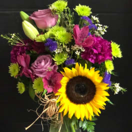 Mixed bouquet with a sunflower, roses, lilies, and purple blooms in a glass vase