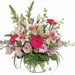 Round glass vase with pink lilies, bright pink gerbera daisies, and pastel blooms in a loose arrangement