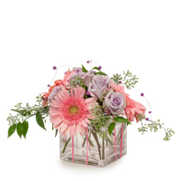 Low pink gerbera daisies and lavender roses in a clear cube vase with decorative beads