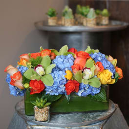 Rectangular floral arrangement with blue hydrangeas, orange and yellow roses, and orchids