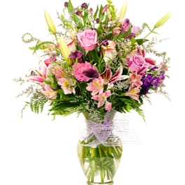 Tall mixed bouquet of pink roses, lilies, and purple accents in a clear glass vase with a mesh bow.