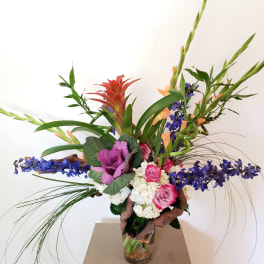 Tall tropical bouquet with pink roses, purple blooms, and a glass vase