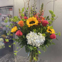 Tall mixed bouquet with sunflowers, roses, and white hydrangea in a glass vase