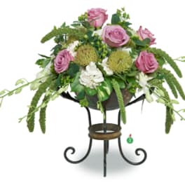 Pink roses, green mums, and white orchids arranged in a dark metal pedestal bowl on a scroll stand