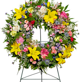 Round standing wreath with yellow lilies and mixed pink, white, and purple flowers on a metal easel.