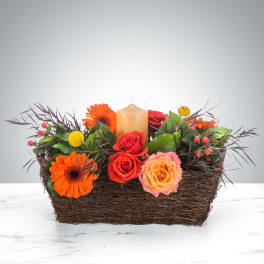 Low basket centerpiece with orange gerbera daisies, red and peach roses, and a cream pillar candle.
