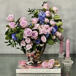 Lavender and pink rose arrangement with blue flowers in a silver vase beside pink candles