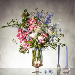 Tall arrangement of pink orchids and blue flowers in a glass vase with two blue taper candles beside it