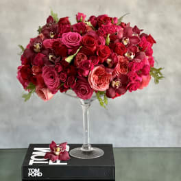 Tall glass compote filled with dense red and hot pink roses and orchids on a black book.
