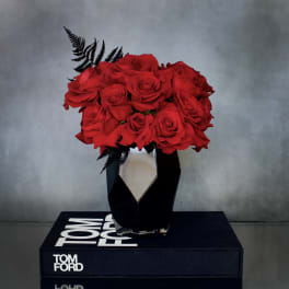 Compact arrangement of red roses with black fern accents in a faceted black vase on a book.