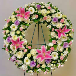 Circular floral wreath with pink lilies, white roses, and purple roses on a stand