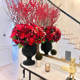 Two large red floral arrangements in black urns on a console table