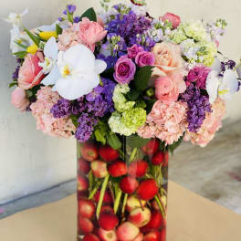 Mixed pink, purple, and white flowers in a tall glass vase filled with apples