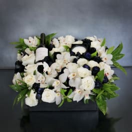 White roses and orchids arranged around a wine bottle and candle in a black box.