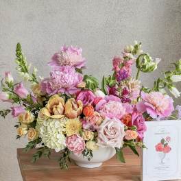 Large pastel flower arrangement in a white bowl with a cookie box beside it.