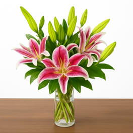 Pink and white lilies with green buds arranged in a clear glass vase on a wooden surface