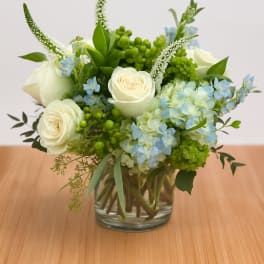 Low round arrangement of white roses, blue hydrangeas, and green accents in a clear glass vase.