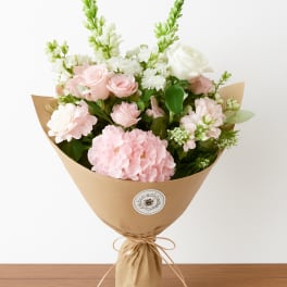 Hand-tied bouquet of soft pink and white flowers wrapped in brown kraft paper.
