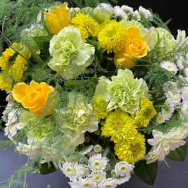 Round arrangement of yellow roses, green carnations, white chrysanthemums, and airy fern-like greenery.