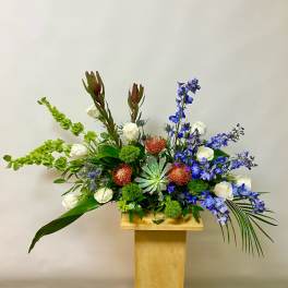 Upright floral arrangement with white roses, blue flowers, and green accents on a wooden pedestal.