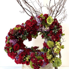 Circular arrangement of red roses, purple blooms, and green succulents with branches in a rustic square container