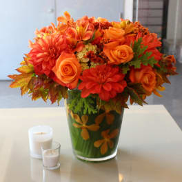 Orange floral arrangement in a glass vase with two candles nearby