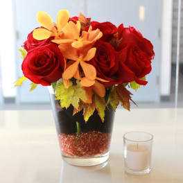 Red roses and orange flowers in a glass vase beside a small candle