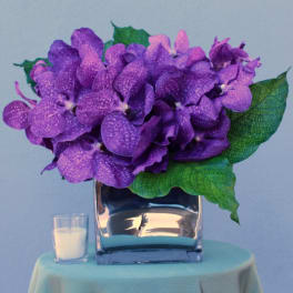 Purple orchid arrangement in a glass vase with a small candle beside it