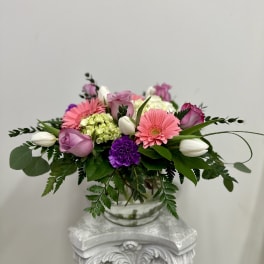 Low glass vase arrangement with pink gerbera daisies, purple roses, white tulips, and green hydrangea