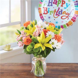 Bouquet of yellow lilies, orange roses, and white daisies in a glass vase with a birthday balloon