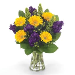 Yellow gerbera daisies and purple flowers in a clear glass vase