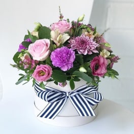 Pink and purple flowers arranged in a white round box with a striped ribbon