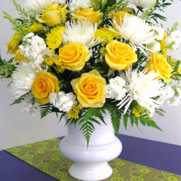 Tall arrangement of yellow roses, white mums, and white blooms in a white urn vase on a patterned runner