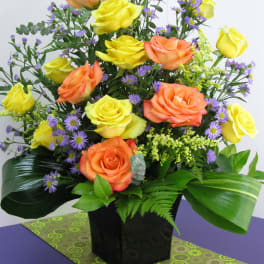 Tall arrangement of yellow and orange roses with small purple flowers in a black cube vase