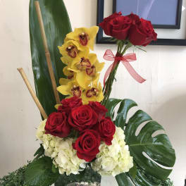 Red roses and yellow orchids arranged in a glass vase with large tropical leaves