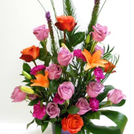 Tall arrangement of lavender roses, orange lilies, and fuchsia blooms in a purple vase with a folded green leaf.