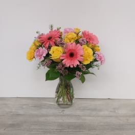 Pink gerbera daisies, yellow roses, and pink carnations arranged in a clear glass vase