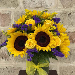Sunflowers, yellow roses, and purple filler flowers in a glass vase with a yellow ribbon.