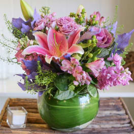 Round arrangement of pink lilies and mixed purple and pink flowers in a green-wrapped glass vase