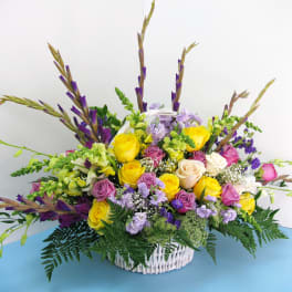 Tall mixed arrangement of yellow and pink roses with purple accents in a white wicker basket