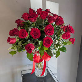 Tall arrangement of red roses with white filler in a clear glass vase tied with a red ribbon.