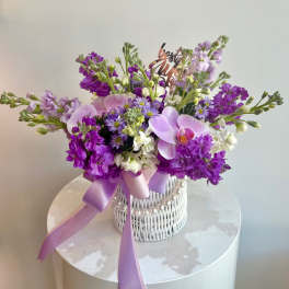 Purple and white floral arrangement in a white wicker basket with a lavender ribbon