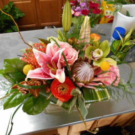Low arrangement of pink lilies, roses, protea, orchids and ranunculus in a rectangular glass vase