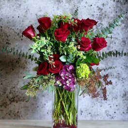 Red roses arranged in a clear glass vase with mixed greenery and purple hydrangea