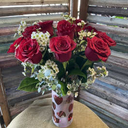 Red roses arranged in a heart-cutout vase with small white filler flowers.