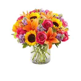 Mixed bouquet of sunflowers, roses, lilies, and purple filler in a glass vase