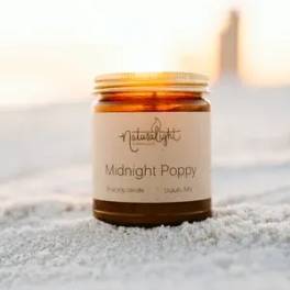 Amber jar candle labeled Midnight Poppy sitting on sand at sunset