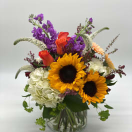 Bouquet of sunflowers, roses, hydrangea, and purple blooms in a glass vase