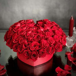 Heart-shaped box of red roses on a reflective surface with scattered petals.