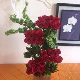 Tall arrangement of red roses and green bells of Ireland in a brown cube vase